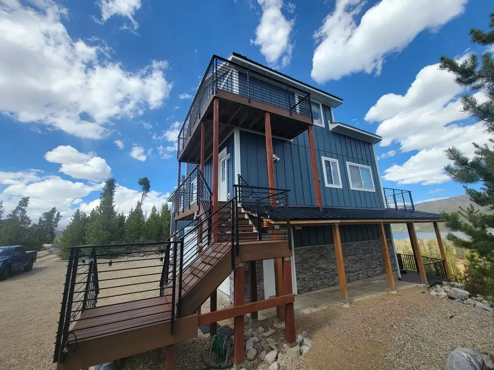 Modern multi-level mountain home with elevated composite decks, black metal railings, and exterior stairs overlooking a lake and pine forest beneath a bright blue sky with scattered clouds.