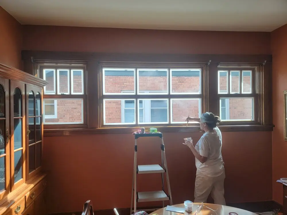 A candid photograph taken from a room corner shows a woman with her grey and brown hair tied up in a bun, wearing a light grey t-shirt and light beige pants, standing on the right side and facing a bank of nine wooden-framed windows as she holds a paint brush and a small, clear paint cup while painting the frame between the central and right window panes. A aluminum step ladder with four steps stands in the center foreground, its top tray holding small paint cans and other supplies. Sunlight streams through the large window cluster, which takes up most of the upper wall and overlooks a brick building with white trim, and casts a warm, soft glow into the room with warm-toned terracotta or rust-orange walls. To the left, a large, ornate wooden hutch or cabinet with curved legs and shelves is visible, with a wooden table in the bottom center foreground and a smaller side table to the right, showing parts of a room ready for painting.
