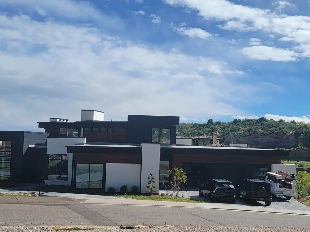 A striking, modern Colorado home exterior featuring a sleek combination of dark charcoal panels, crisp white stucco, and warm wood cladding. The installation by The Install Company includes multiple sleek garage doors and large black-framed windows, all set against a rocky hill and a blue, cloudy sky. Several SUVs are parked on the large concrete driveway.
