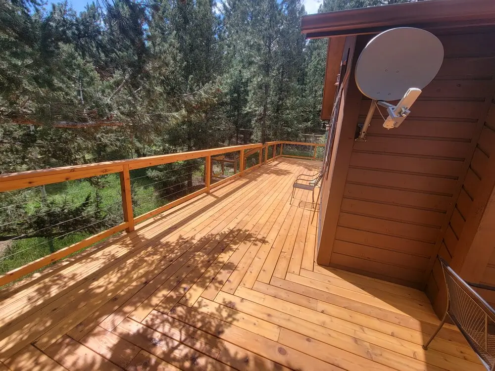 Freshly stained wood deck with cable railing overlooking a peaceful pine forest, attached to a mountain home with a spacious outdoor living area and scenic wooded views.