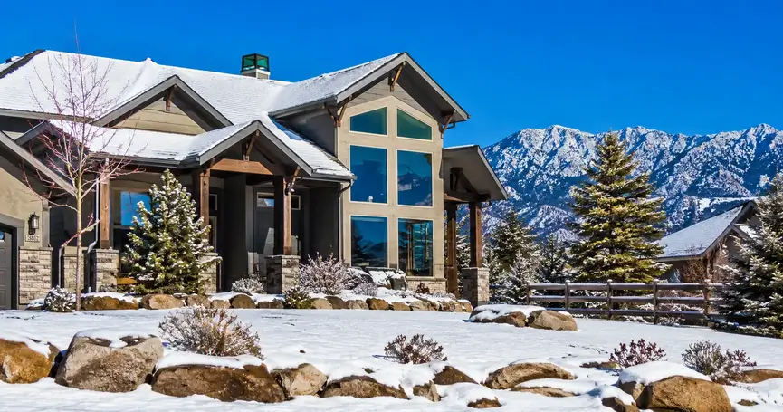 Modern Front Range Colorado home with large floor-to-ceiling windows, a snow-dusted yard, evergreen trees, and snow-covered mountains beneath a bright blue winter sky.